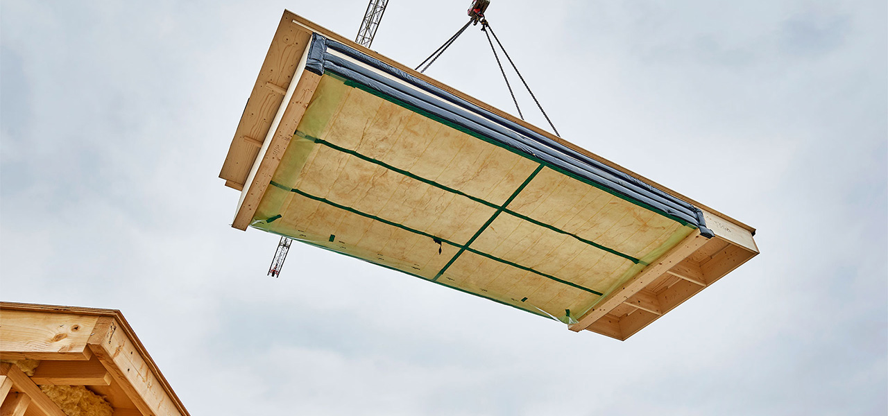 Een grote constructieve multiplex plaat wordt met behulp van een kraan opgetild en naar zijn plaats gebracht. De plaat is voorzien van isolatiemateriaal en versterkingen, die zichtbaar zijn aan de onderkant van de constructie.