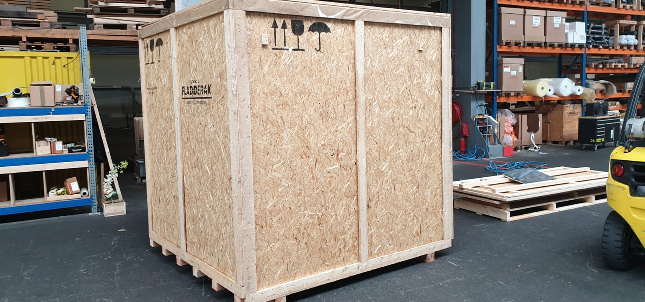 Een grote houten transportkist gemaakt van OSB-platen, voorzien van markeringen en labels, in een magazijnomgeving met stapels pallets en gereedschap.