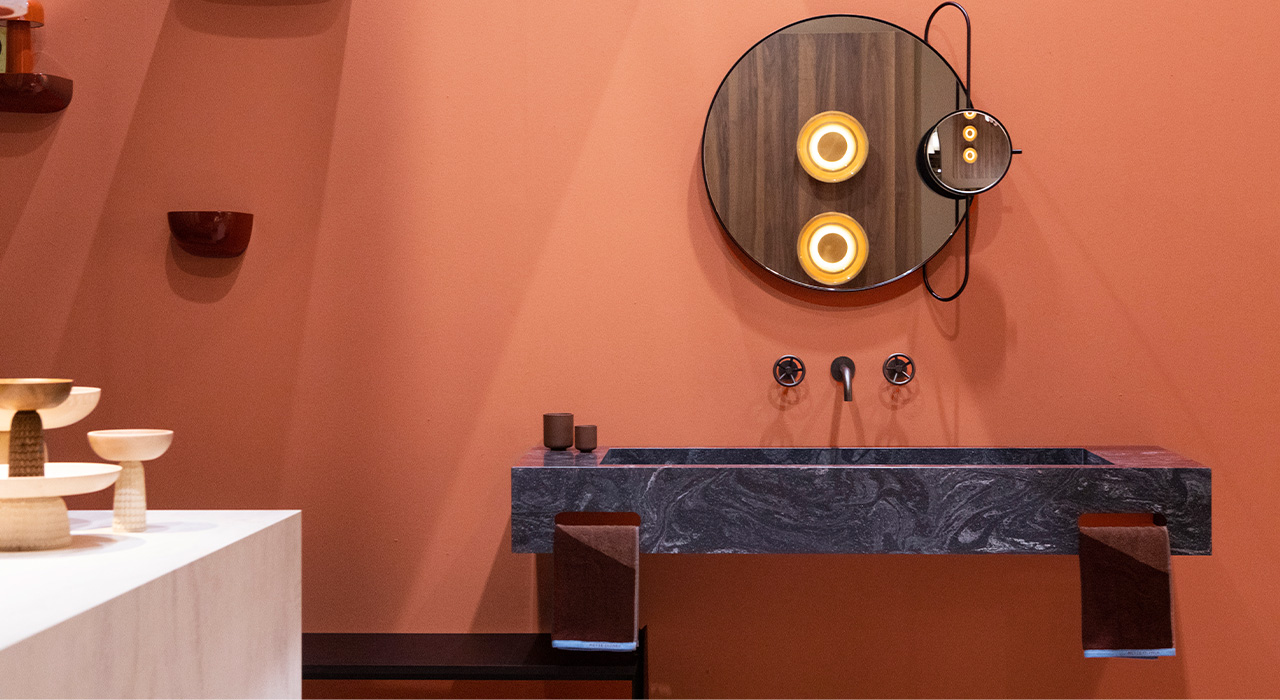 Leeuwerik Plaat stand Design District 2 – Marmeren wastafel met ronde spiegel en designverlichting op een warme terracotta muur.