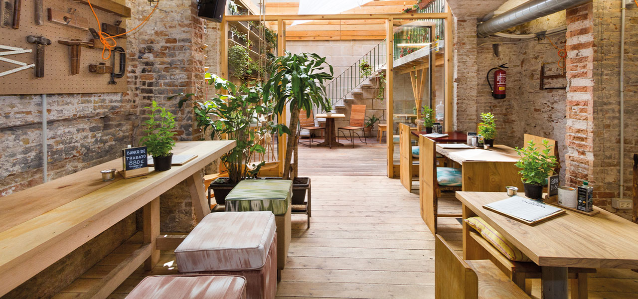 Een sfeervol interieur van een rustieke werkplaats of eetruimte, met houten meubels, planten en gereedschappen tegen een bakstenen muur.