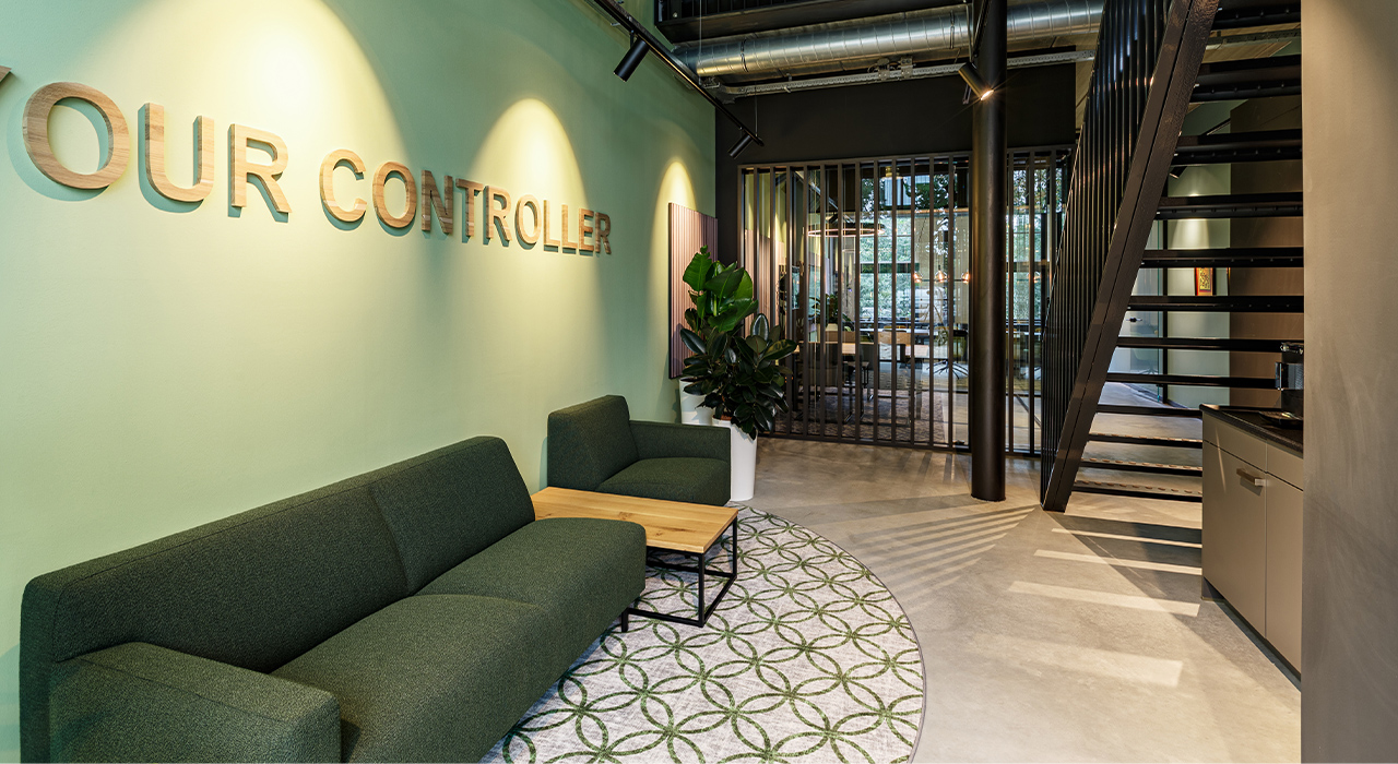Your Controller Vught 1 – Wacht- en ontvangsthoek met groene bank, houten tafel en industriële trap.