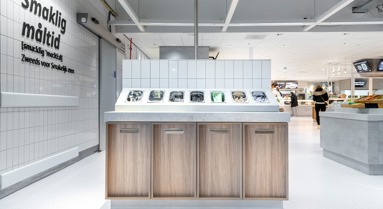 Zelfbedieningsstation met houten kasten en ingebouwde bestekbakken, voor een witte betegelde muur met "smaklig måltid."